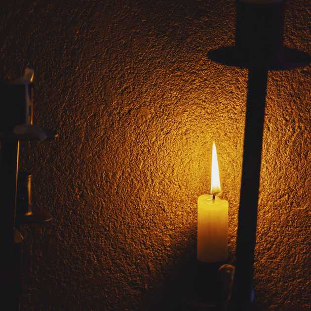 A burning candle casting its yellow light on a rugged wall and 3 empty candleholders